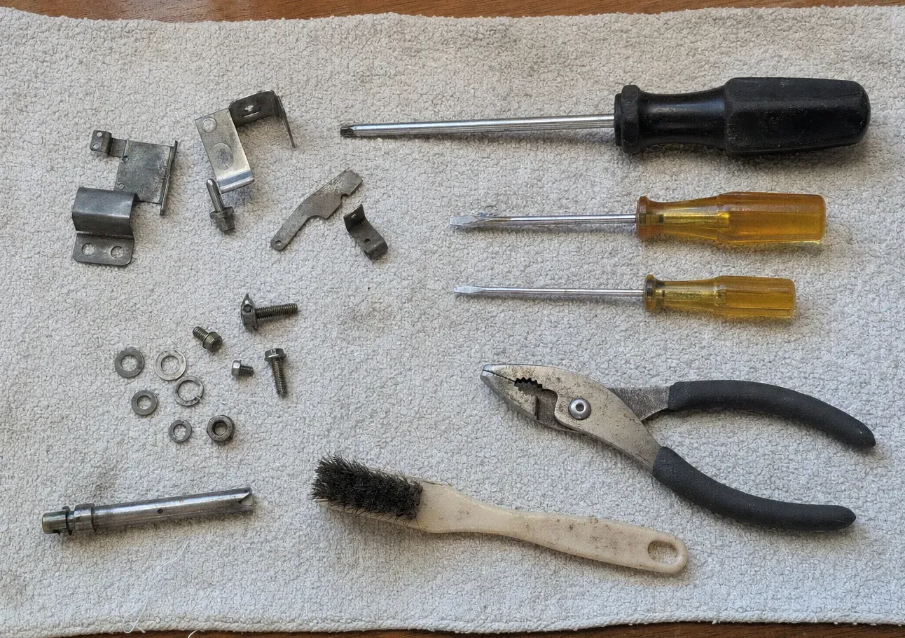 Oven door hinge plates, amber-handled screwdrivers, snips and a wire brush laid out with bolts and washers